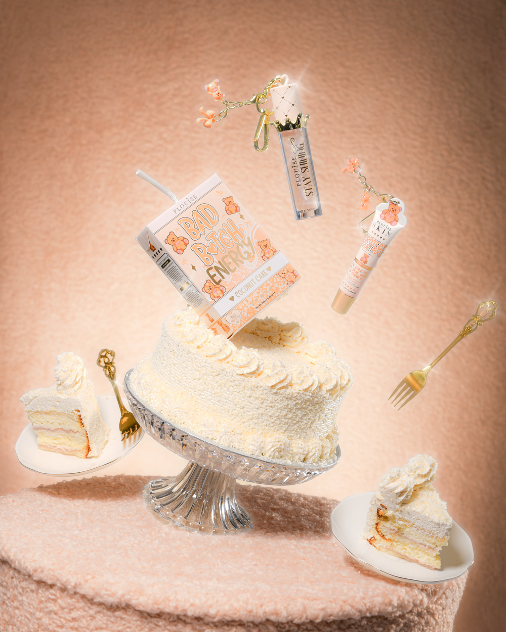 Een taart op een tafel van teddystof met een P.Louise Teddy Glitter Bad Bitch Energy Lip Duo in kokoscake met daarin een lipconditioner en lipolie die rond de taart zweeft