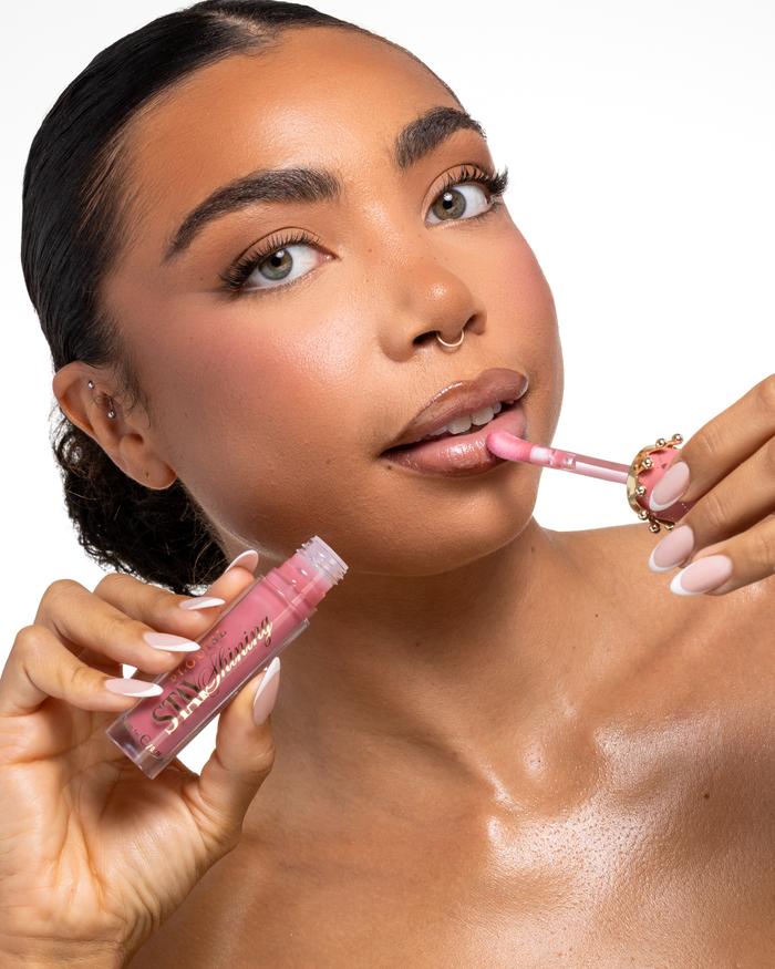 Un gros plan d'une femme appliquant un produit liquide pour les lèvres rose P.Louise Stay Shining Lip Oil avec un applicateur pied de biche tout en tenant le tube dans son autre main, sur un fond blanc propre. arrière-plan.