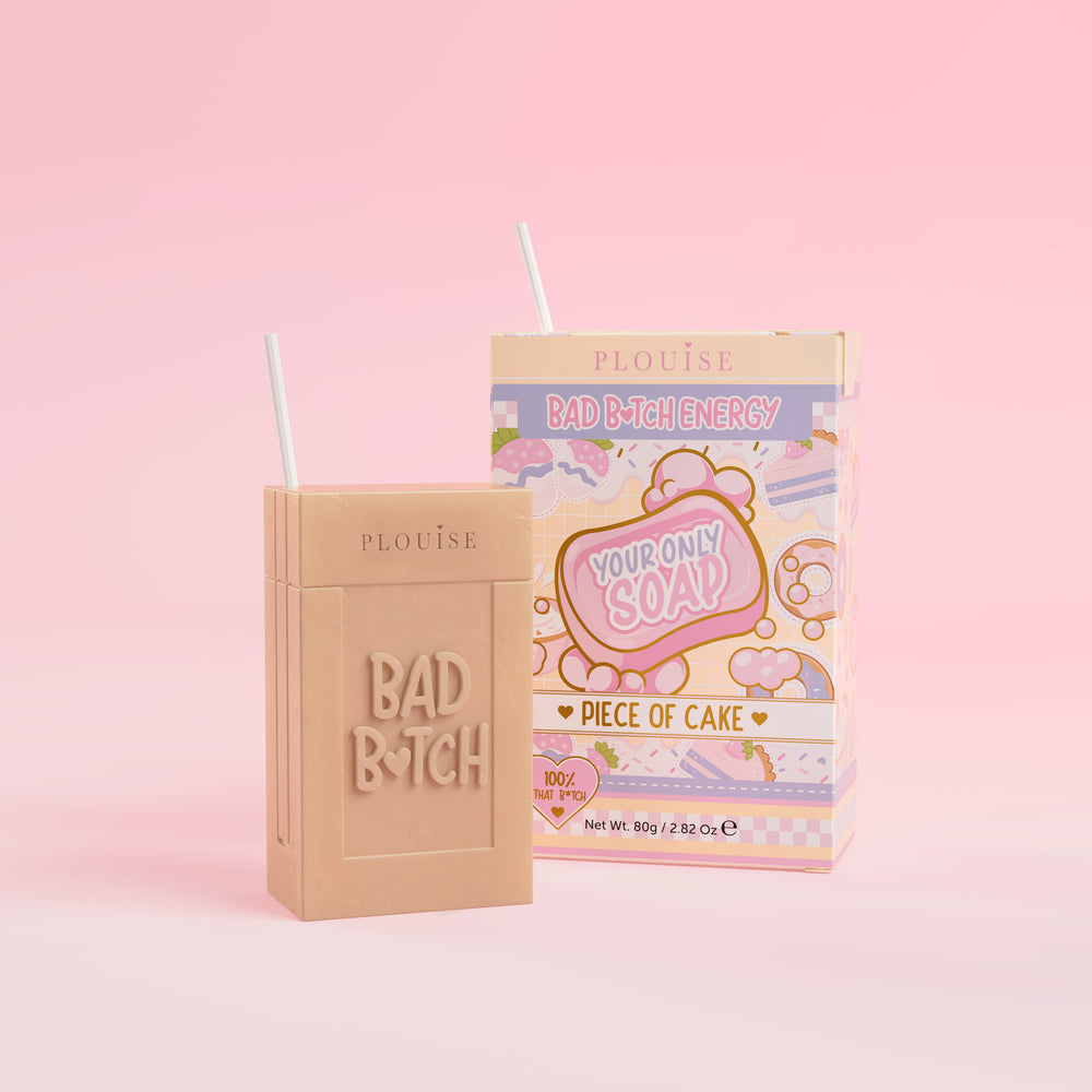 Een beige stuk zeep met 'Bad Bitch' erop gegraveerd, vergezeld van een kleurrijk sapdoosje met het opschrift 'Piece of Cake' erop, gepresenteerd op een roze achtergrond.