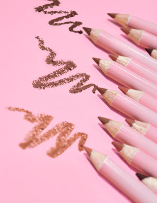 A collection of P.Louise ultimate definition sharpenable lip liners in a variety of nude shades, laid out on a pink background with some lines of the product drawn on the surface.